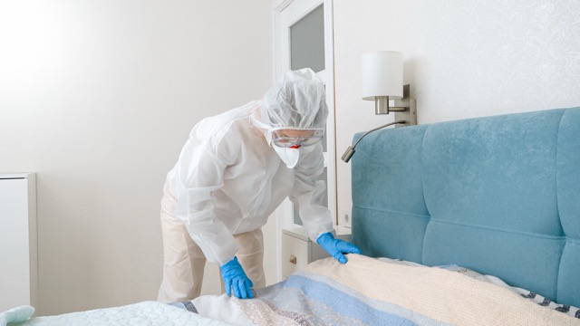Young housewife in protective biohazard suit making up bed during covid-19 pandemic and lockdown