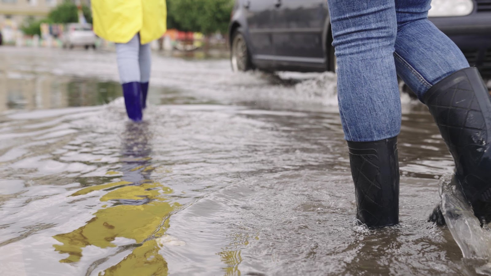 Two individuals in rain boots navigating a flooded street amidst flood damage and mold