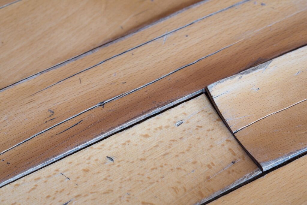 Close-up of warped and peeling light wood floor planks showing water damage.