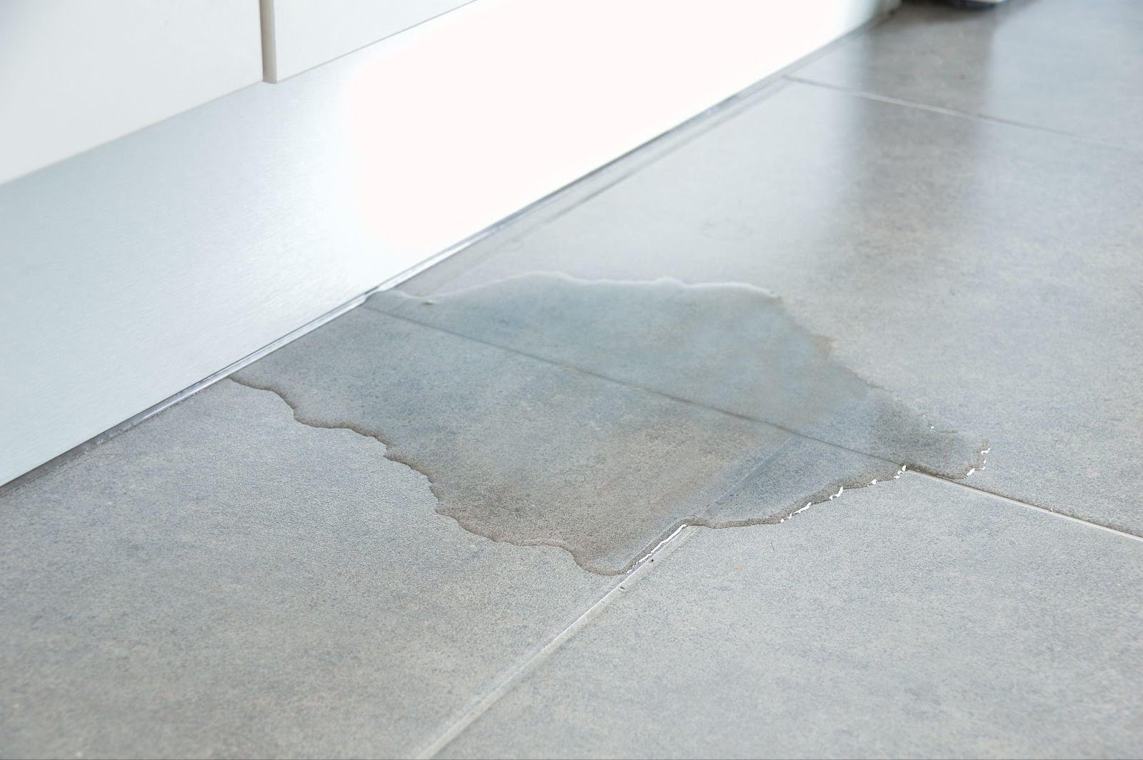 A clear water puddle reflecting light on the tiled floor of a kitchen, surrounded by kitchen appliances and furniture