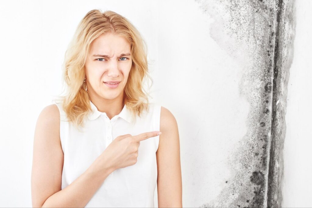 A woman in a white top pointing at mold on a wall with a concerned expression.