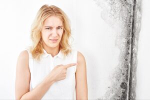 A woman in a white top pointing at mold on a wall with a concerned expression.