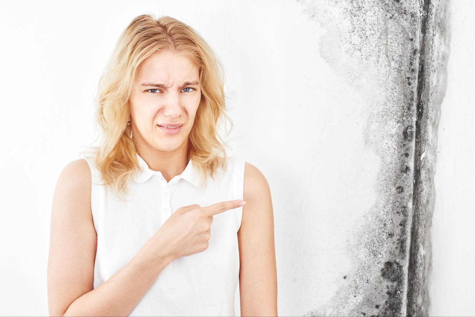 A woman in a white top pointing at mold on a wall with a concerned expression.