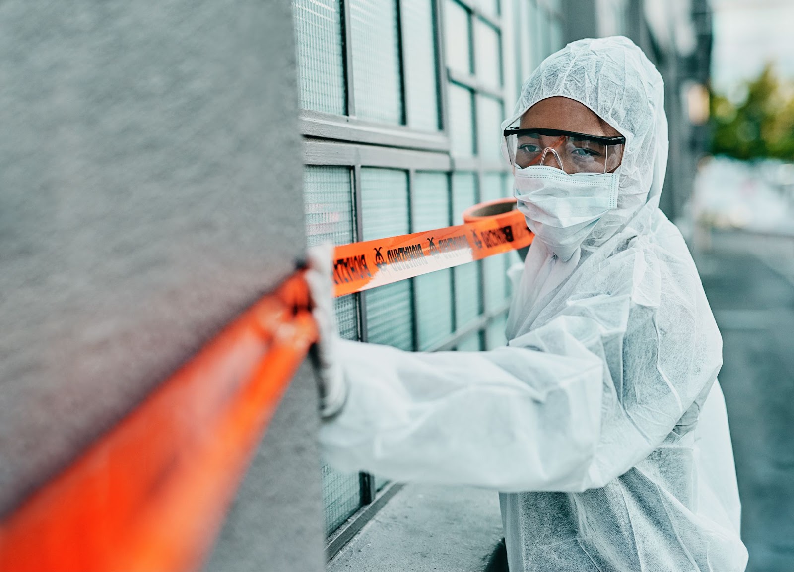 Person in a white hazmat suit with goggles and mask, standing behind orange caution tape outside a building.