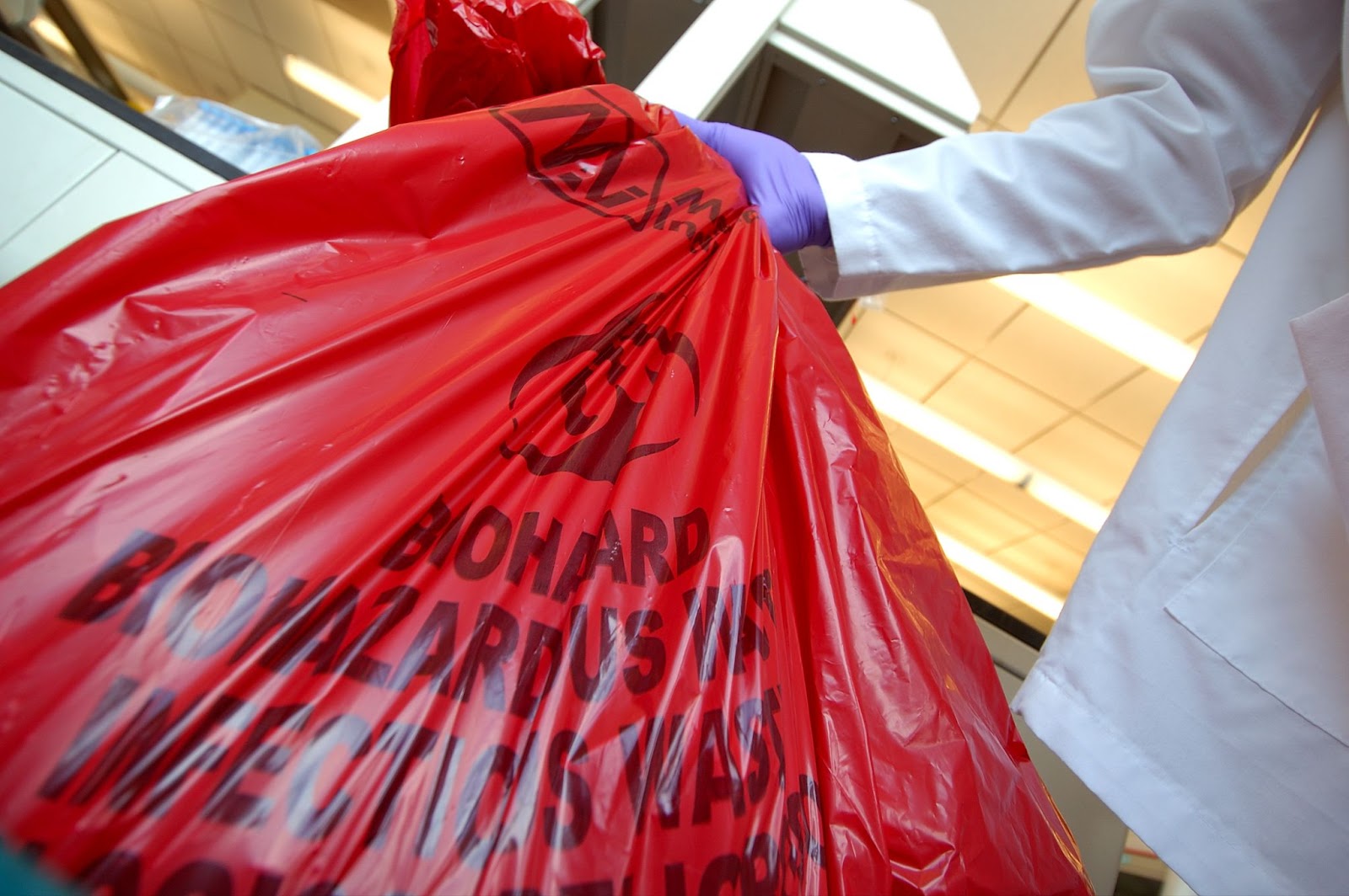 Person in a white lab coat and purple gloves holding a red biohazard waste bag labeled "BIOHAZARDOUS WASTE."