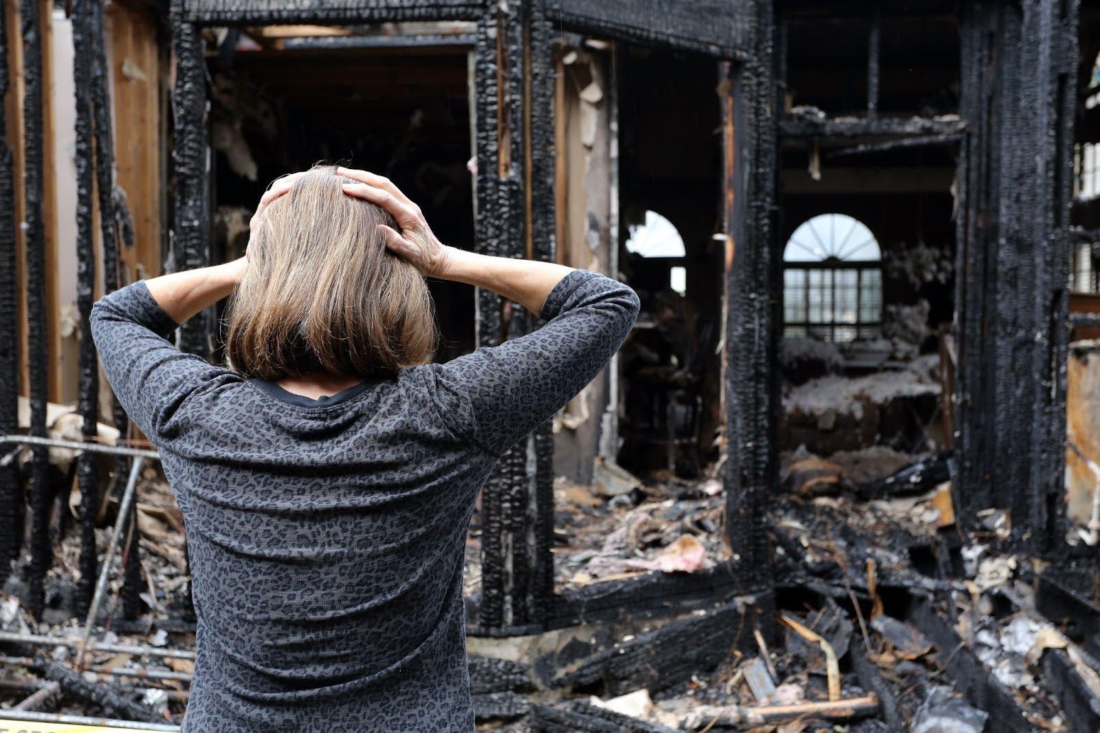 A woman stands solemnly in front of a charred house reflecting on the aftermath of a fire