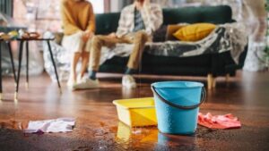 A blue bucket and yellow tray collecting water leaking from a ceiling onto a floor, with a couple sitting on a couch in the background.