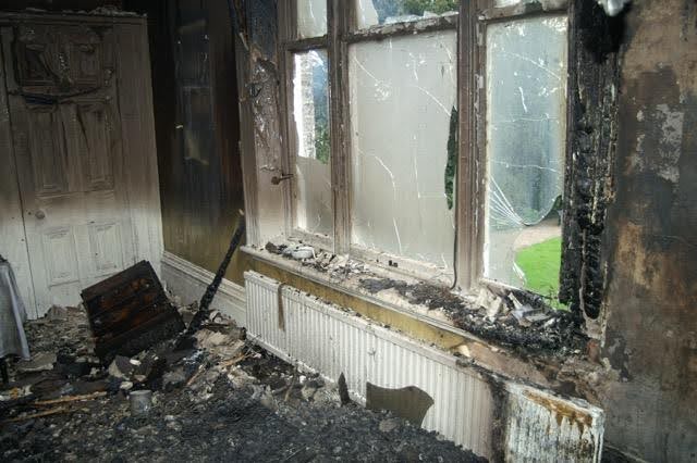 A charred room with a broken window and an open door showcasing the aftermath of a fire