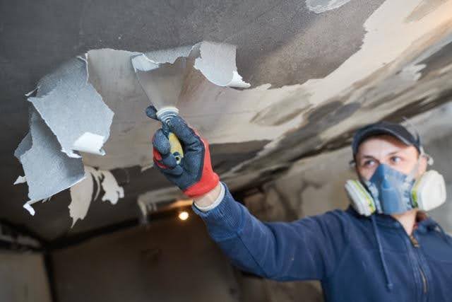 A man in a blue shirt and mask is applying paint to the ceiling with a roller