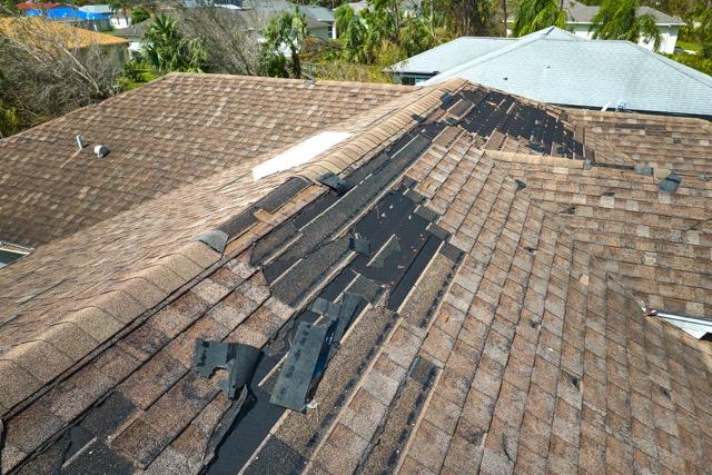 Aerial view of a brown shingle roof with significant wind damage and missing shingles after a storm.