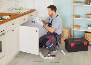 A plumber in work overalls kneeling in a kitchen, holding a white plastic pipe fitting while repairing the plumbing under a sink.