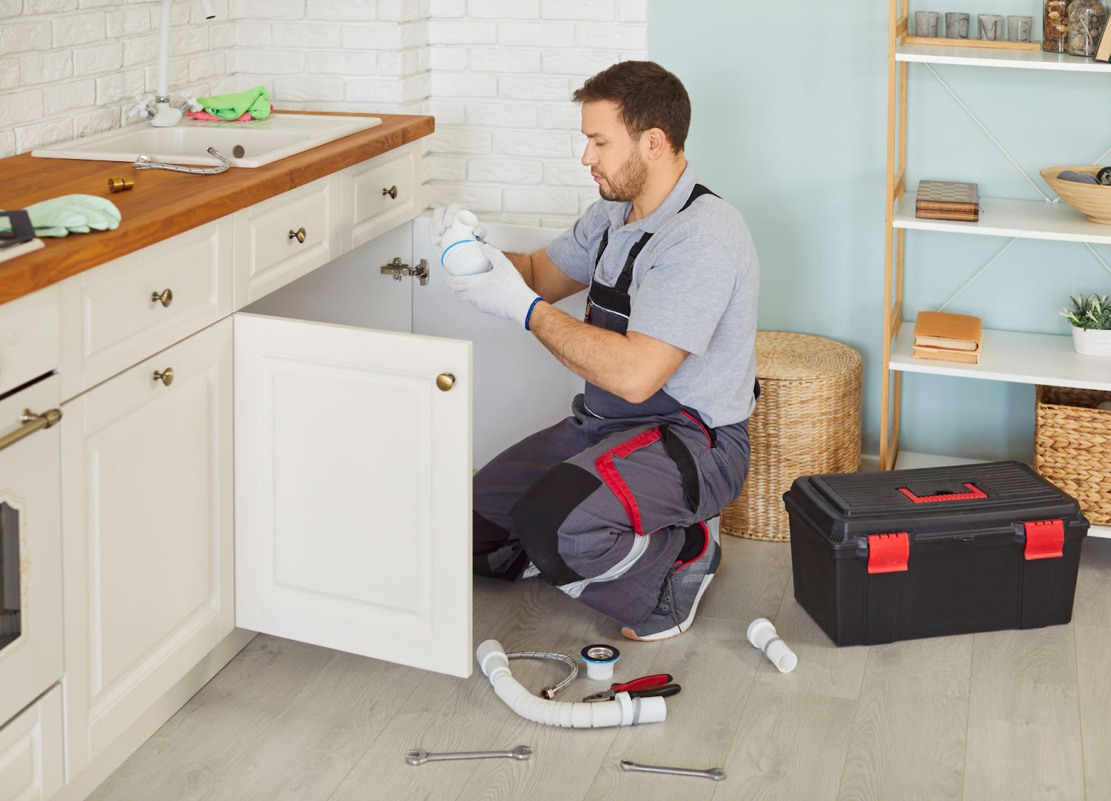 A plumber in work overalls kneeling in a kitchen, holding a white plastic pipe fitting while repairing the plumbing under a sink.