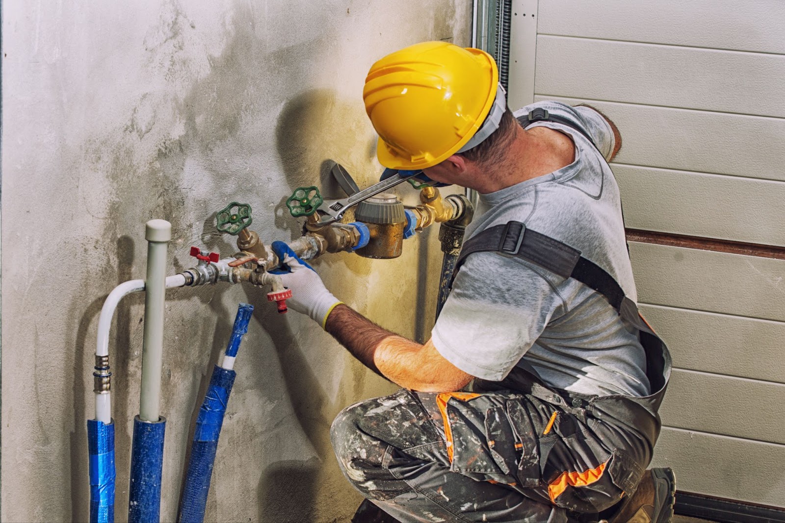 A plumber wearing a yellow hard hat and work overalls uses a wrench to install or repair a water meter and pipe assembly on a concrete wall.