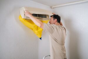 A technician opening the front panel of a wall-mounted ductless mini-split air conditioner, which is fitted with a yellow waterproof cleaning cover to catch runoff.