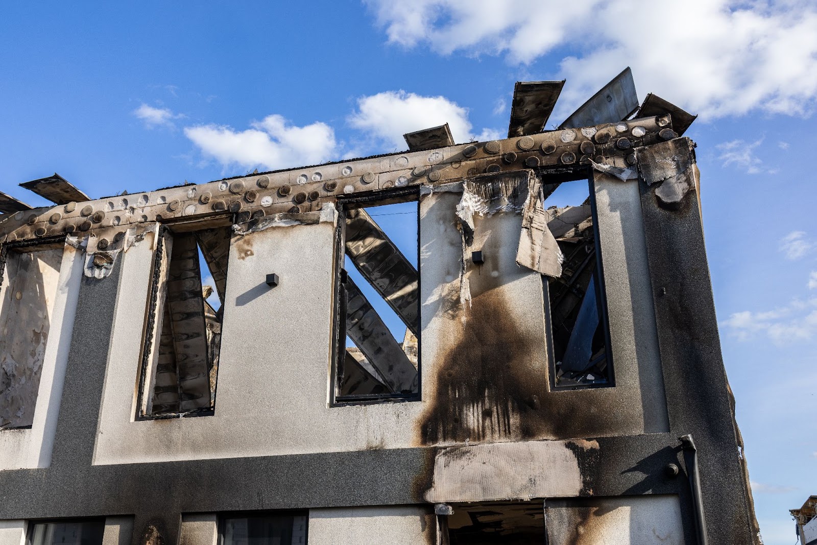 A view of a two-story building with severe fire damage, including charred window frames, a partially collapsed roof, and soot-blackened exterior walls.