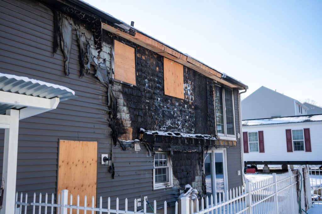 The exterior of a two-story home shows significant fire damage, with charred siding, melted insulation, and several windows boarded up with plywood.