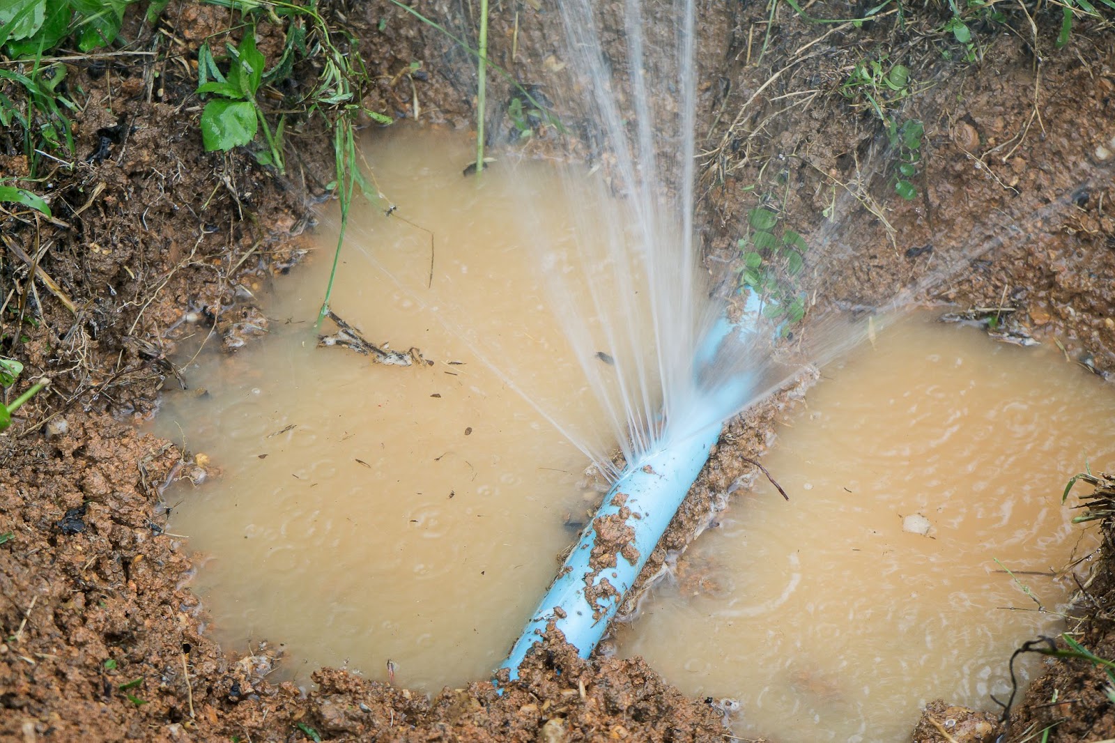 Water spraying high into the air from a burst blue PVC underground water pipe in a muddy excavation hole.