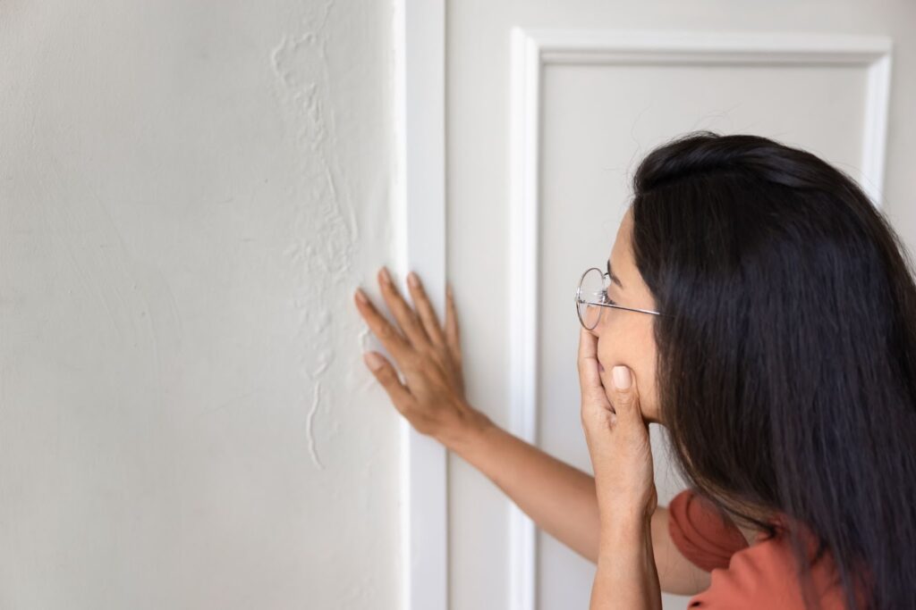 A concerned woman touching a wall with visible water damage and bubbling paint.