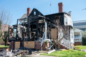 A two-story house severely damaged by fire, showing charred wooden framing, a collapsed roof section, and debris scattered across the yard.