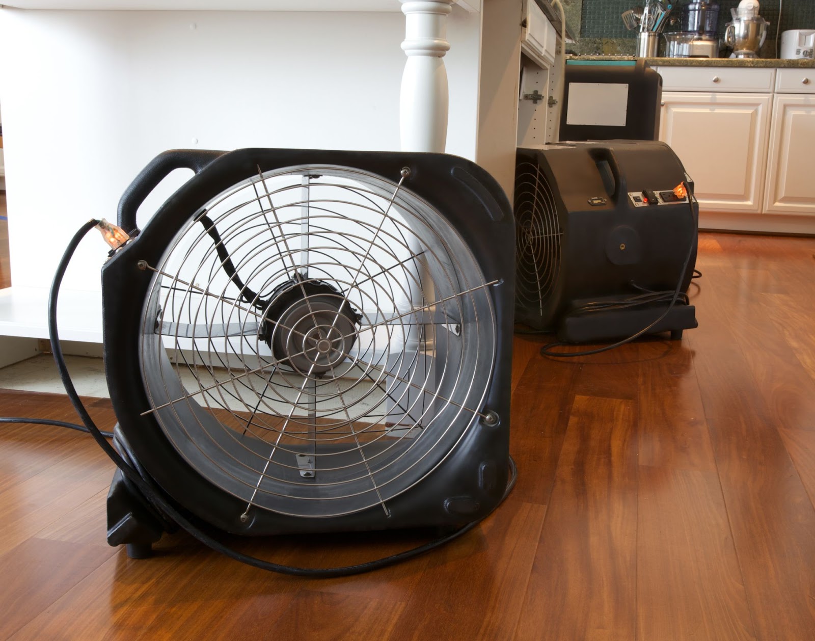 Two industrial air movers placed on a wooden floor in a kitchen to dry out water damage.