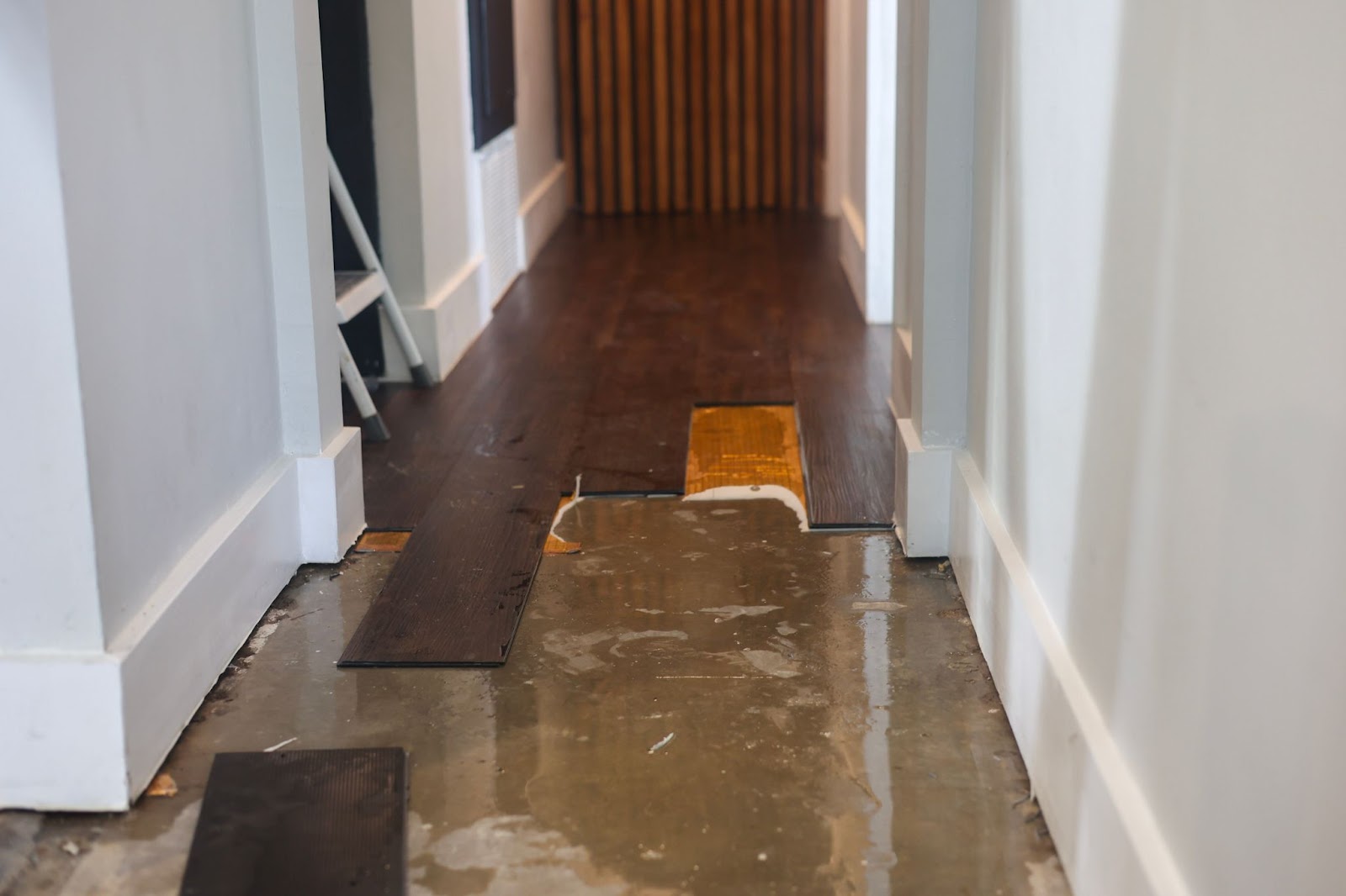 Water damaged dark wood laminate flooring being pulled up to reveal a wet concrete subfloor in a hallway.