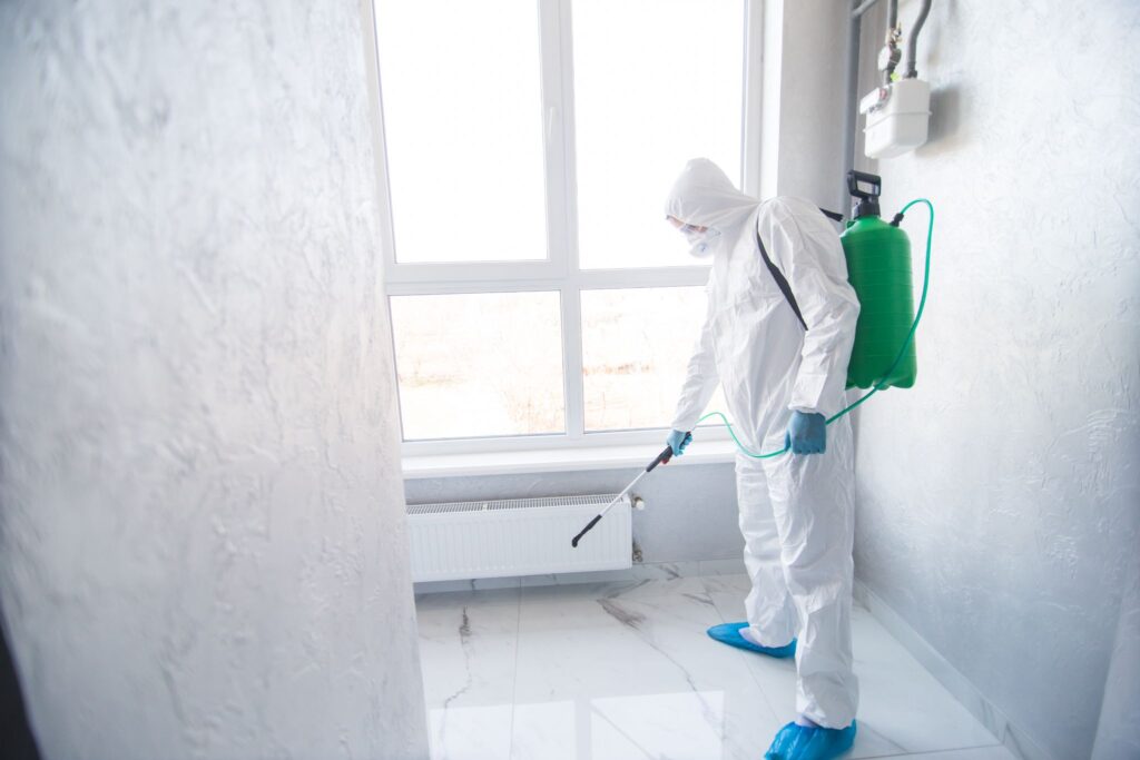 A person in a white hazmat suit and protective gear spraying a floor using a green backpack sprayer.