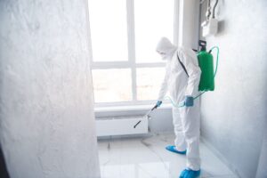 A person in a white hazmat suit and protective gear spraying a floor using a green backpack sprayer.