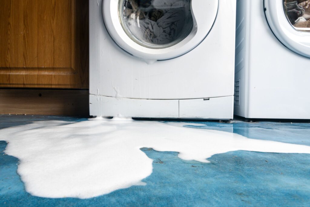 A leaking washing machine overflowing with soap suds onto a blue floor.