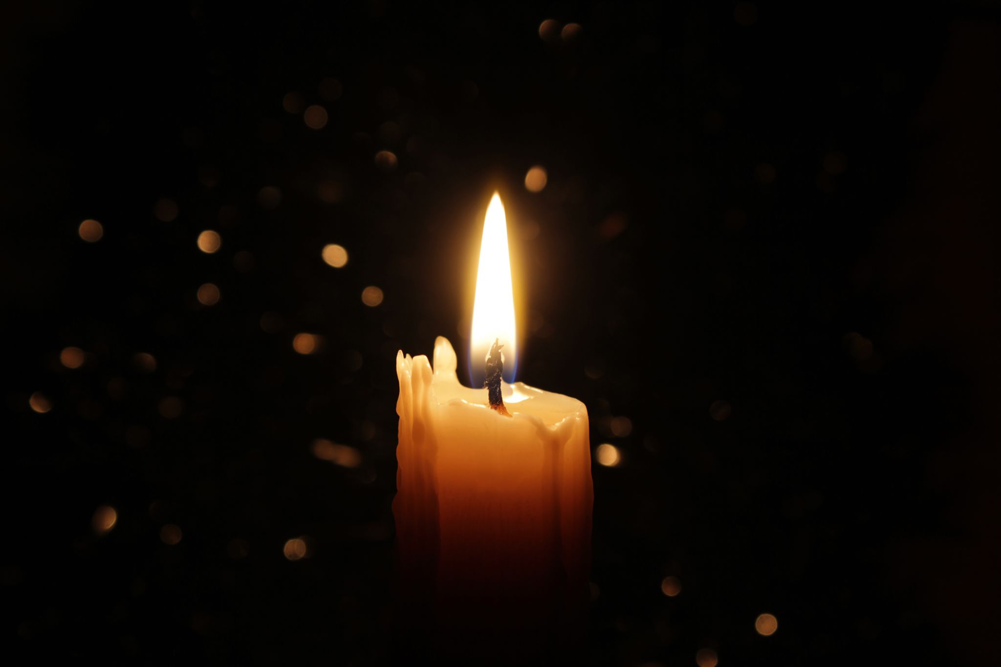 A single lit wax candle burning with a steady flame against a dark background with soft bokeh lights.