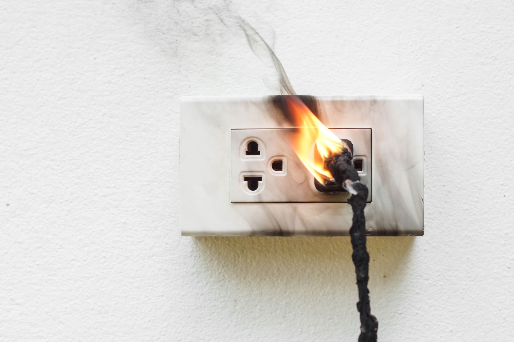 An electrical outlet on a white wall catching fire and emitting black smoke from a plugged-in cord.