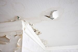 Severe water damage on a white ceiling and wall showing peeling paint, water stains, and a cracked hole.