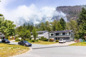 Suburban homes and parked cars sit beneath forested mountains veiled in drifting smoke