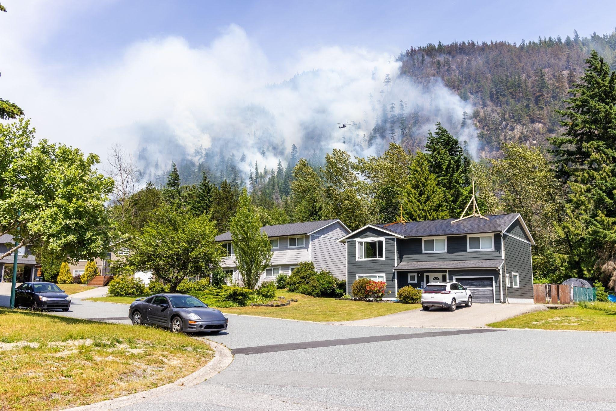 Suburban homes and parked cars sit beneath forested mountains veiled in drifting smoke