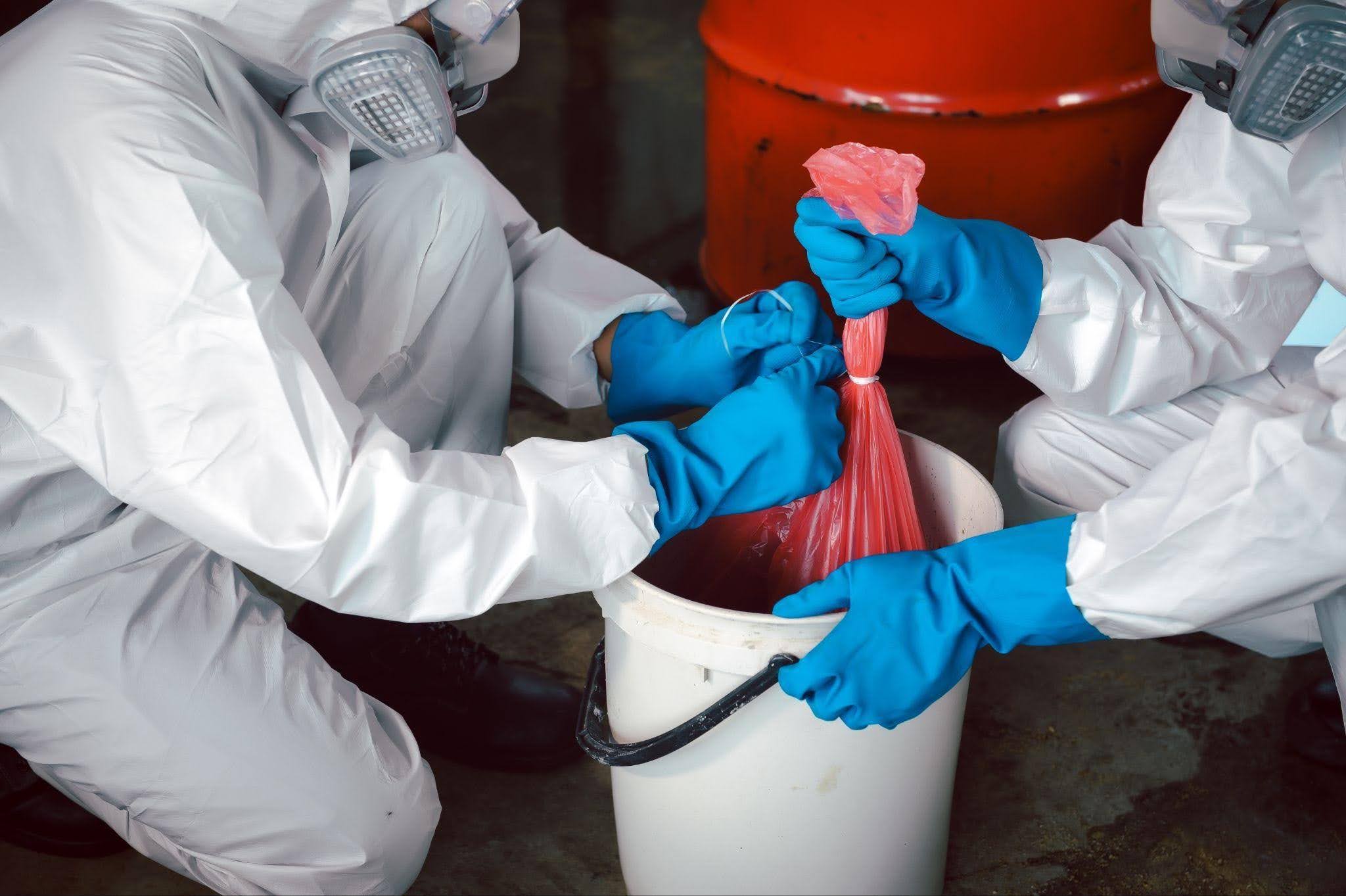 Workers in white hazmat suits and respirators handle a tied red biohazard bag over a white bucket