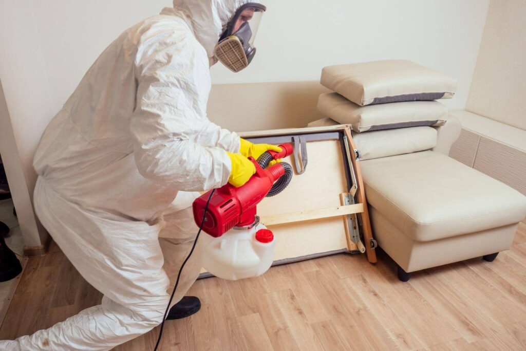 A man in a white suit and protective gear sprays a couch with a cleaning solution