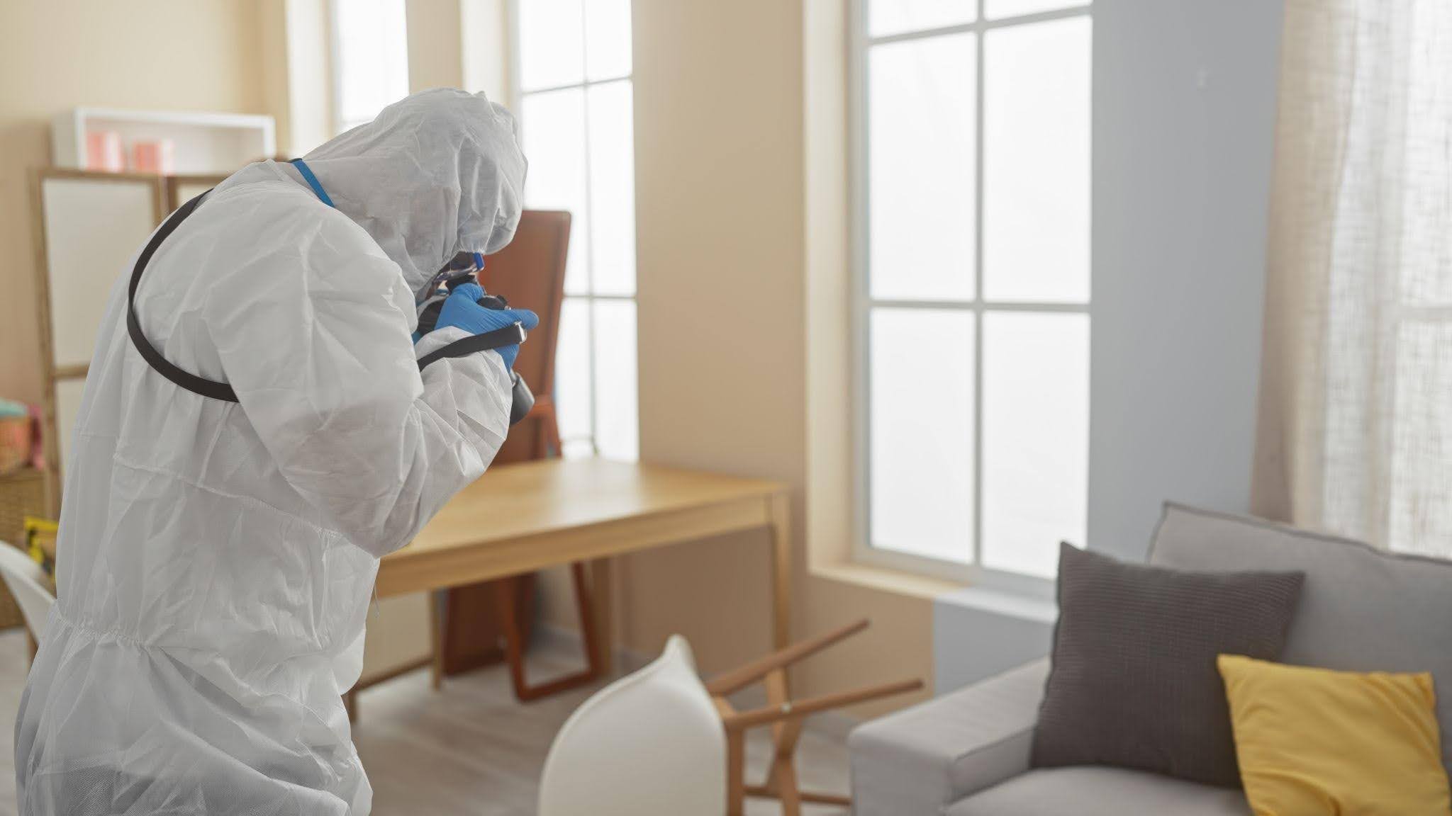 A person in a white protective suit stands in a living room surrounded by furniture and decor