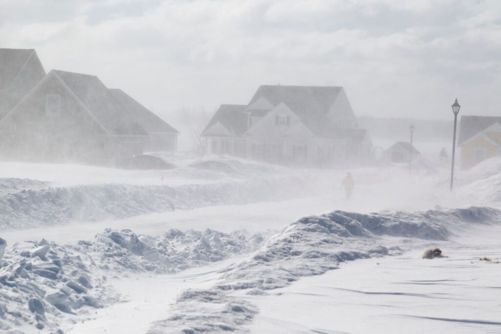 Suburban neighborhood street during a heavy winter blizzard with thick snowbanks and blowing snow.